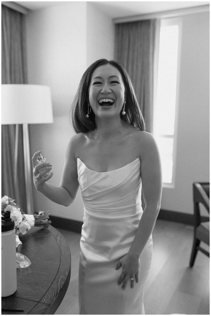 Black and white image of bride laughing while she puts on perfume.