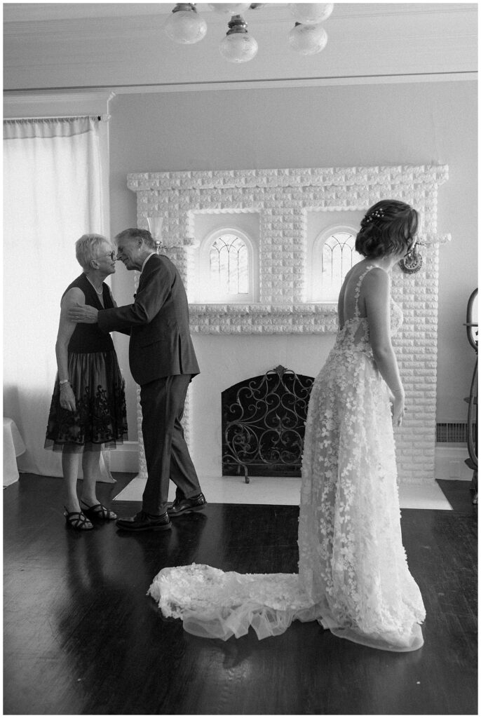 Black and white image of bride's parents hugging while the bride laughs in the foreground.